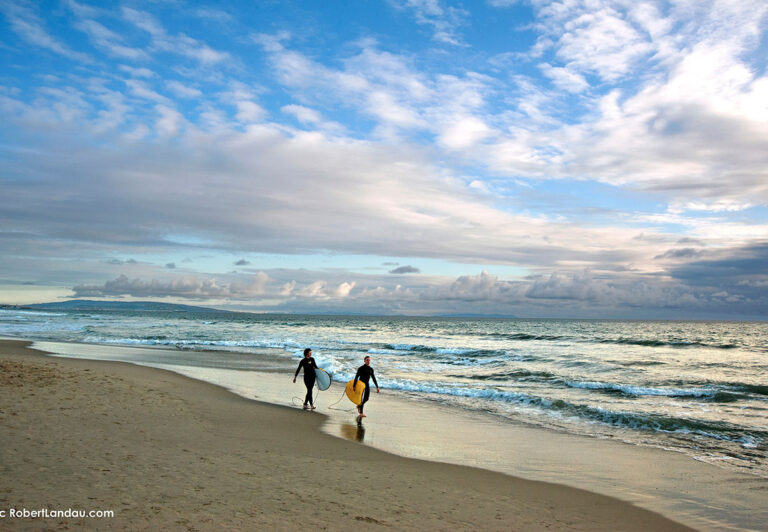 A dramatic sky near day’s end forms the backdrop for a couple of young surfers in wetsuits walking towards Malibu, perhaps in search of that one last wave.