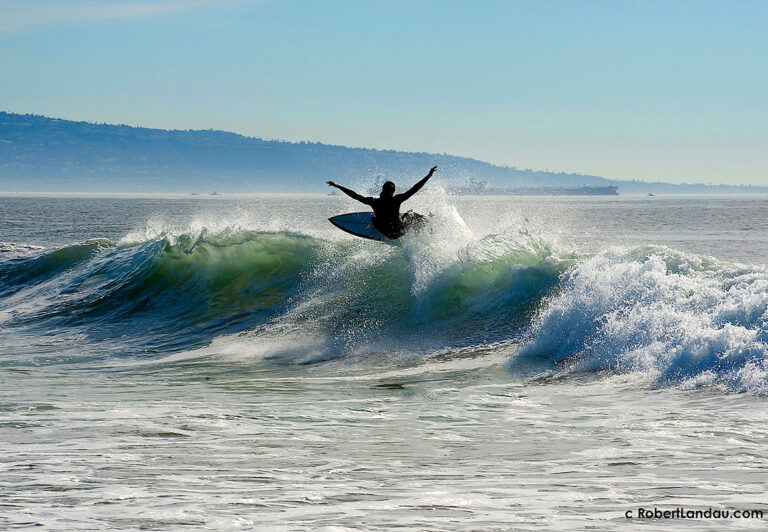 A surfer enjoys the ride on the crest of a breaking wave at Venice Beach with the Palos Verdes Peninsula seen in the background.