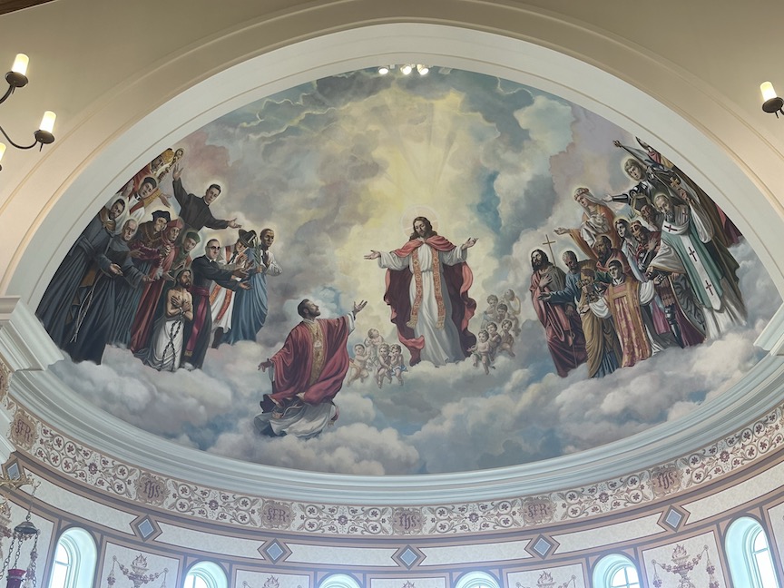Mural appearing on the dome of the roof at the Blessed Stanley Rother Shrine in Oklahoma City introduces Catholic pilgrims to Christian martyrs killed for practicing their faith. Mural shows Stanley Rother in red robe kneeling before Jesus Christ while other martyrs look on.