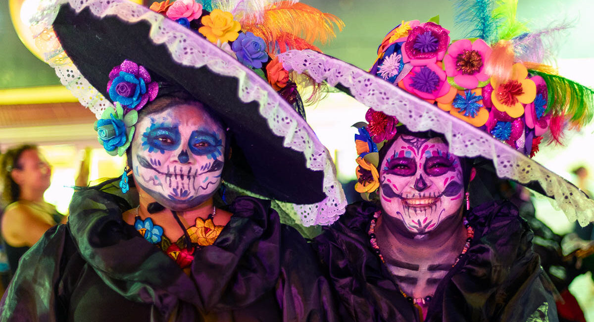 (1) Homepage Photo ISLA_0872 Two happy ladies dressed as La Calavera Catrina. The costume is based on Jose Guadeloupe Posada caricature of a woman skeleton covering up her Indigenous heritage in lots of make-up to make her skin look whiter along with a French dress and fancy hat.