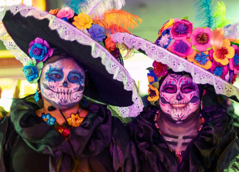 Two happy ladies dressed as La Calavera Catrina. The costume is based on Jose Guadeloupe Posada caricature of a woman skeleton covering up her Indigenous heritage in lots of make-up to make her skin look whiter along with a French dress and fancy hat.