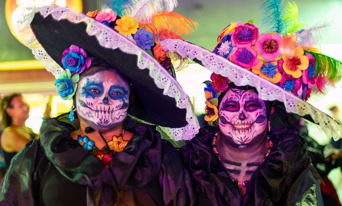 Two happy ladies dressed as La Calavera Catrina. The costume is based on Jose Guadeloupe Posada caricature of a woman skeleton covering up her Indigenous heritage in lots of make-up to make her skin look whiter along with a French dress and fancy hat.