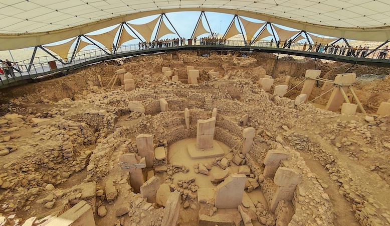 A large canopy covers the Gobekli Tepe site, with visitors looking down from ramps. 