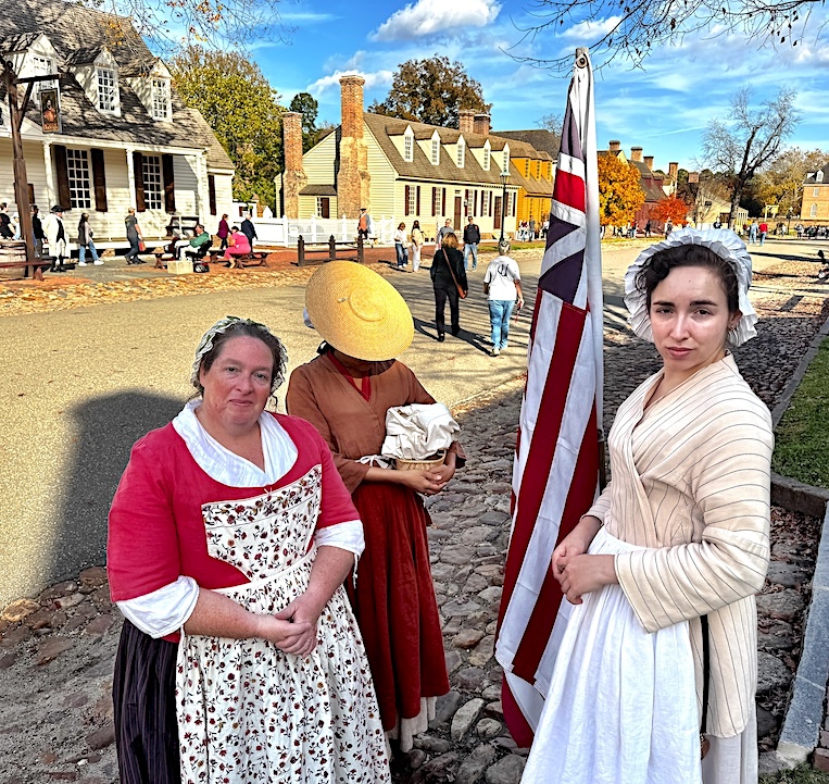 Three working women, one of them an indentured "convict servant" deported from England after being convicted of theft, discuss colonial news while standing on a Gloucester Street sidewalk.
